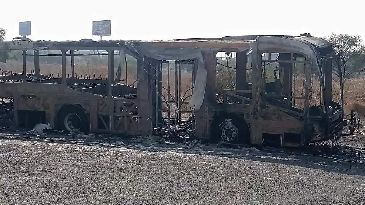 Two charred, burnt-out buses on a barren ground with buildings visible in the background