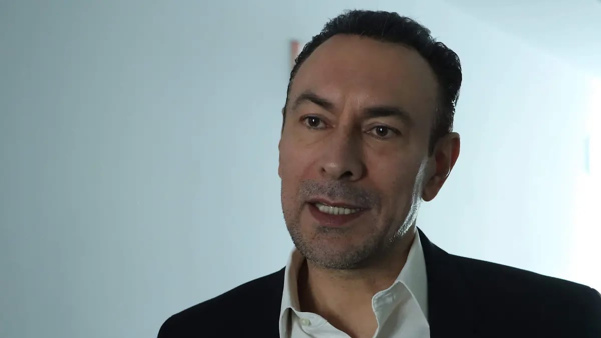 Man in black blazer and white shirt smiling at camera against light blue background