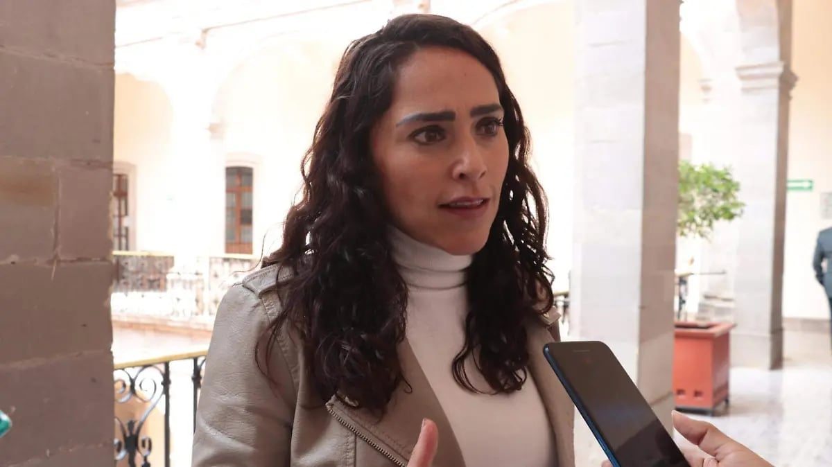 Woman with long dark hair wearing a white turtleneck and brown blazer standing in a colonnade
