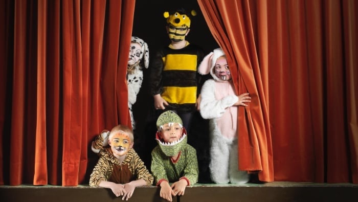 Children in costumes performing on a puppet theater stage with red curtains, including a bee character and other masked figures