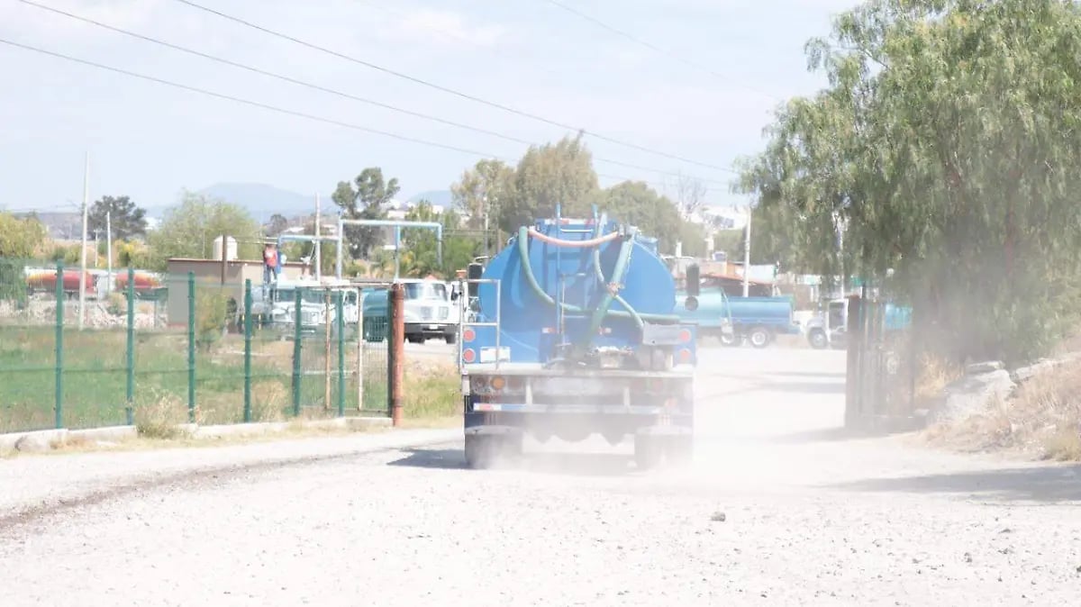 Blue water tanker truck driving on dusty gravel road past playground and houses