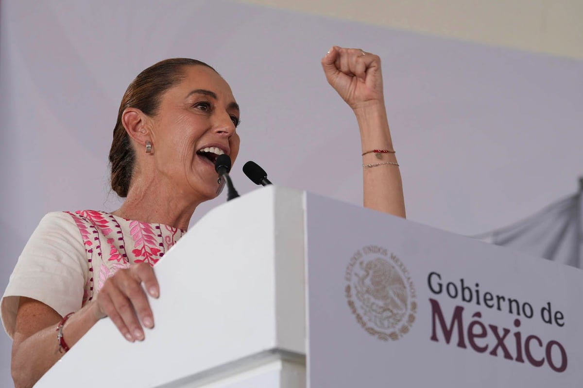 Woman speaking at podium with Gobierno de Mexico logo, raising fist while holding microphone with animated expression
